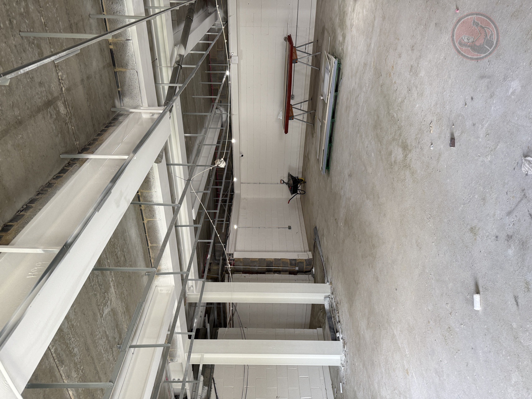 Empty unfinished industrial space with concrete floor, exposed ceiling, white painted walls, a wheelbarrow, and a workbench with materials.