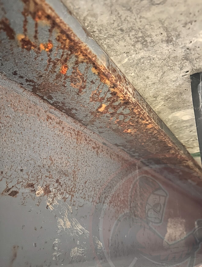 Close-up of a rusty metal surface with peeling paint and corrosion under a concrete structure.