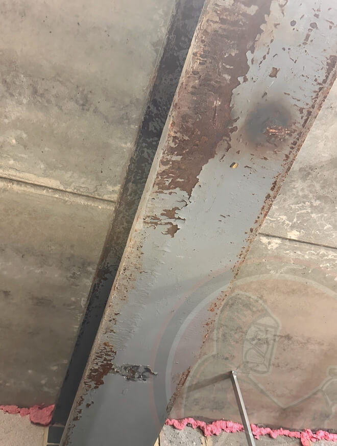 Rusty and peeling painted steel beam on a concrete ceiling with pink insulation visible at the bottom.