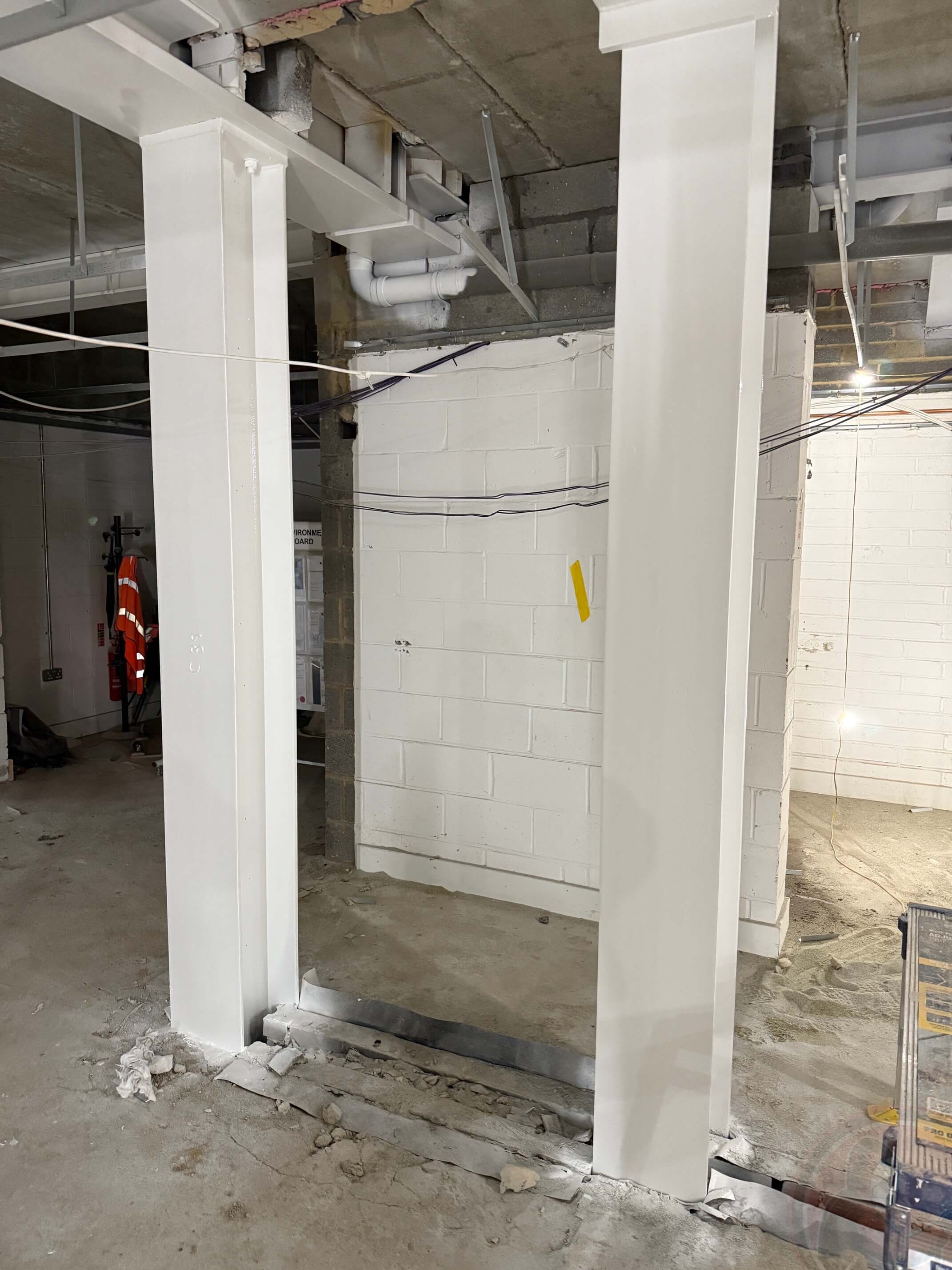 White steel support columns installed in an unfinished basement or industrial space with concrete floor and block walls.