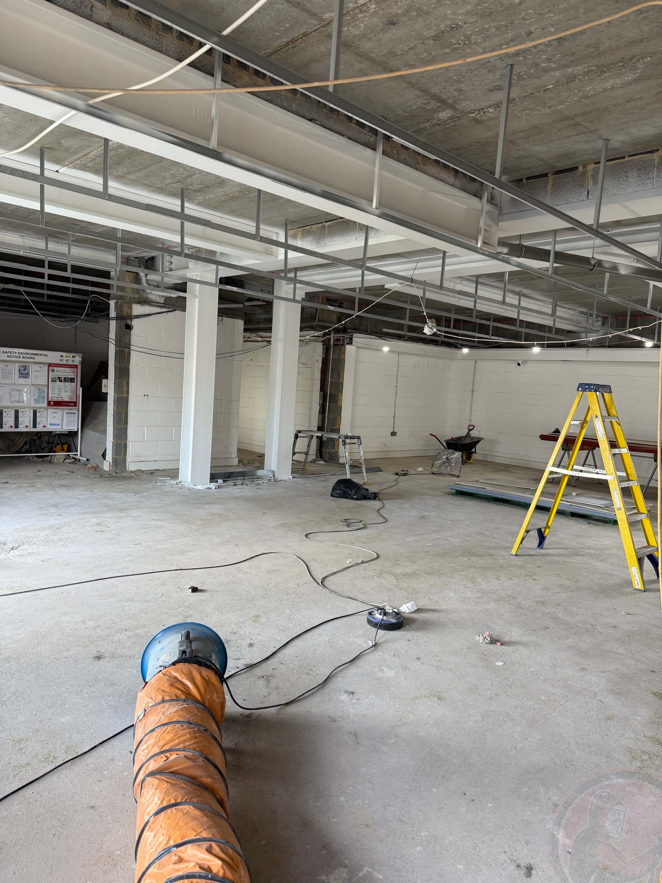 Empty indoor construction site with concrete floor, white pillars, yellow ladder, ventilation duct, and construction tools scattered around.