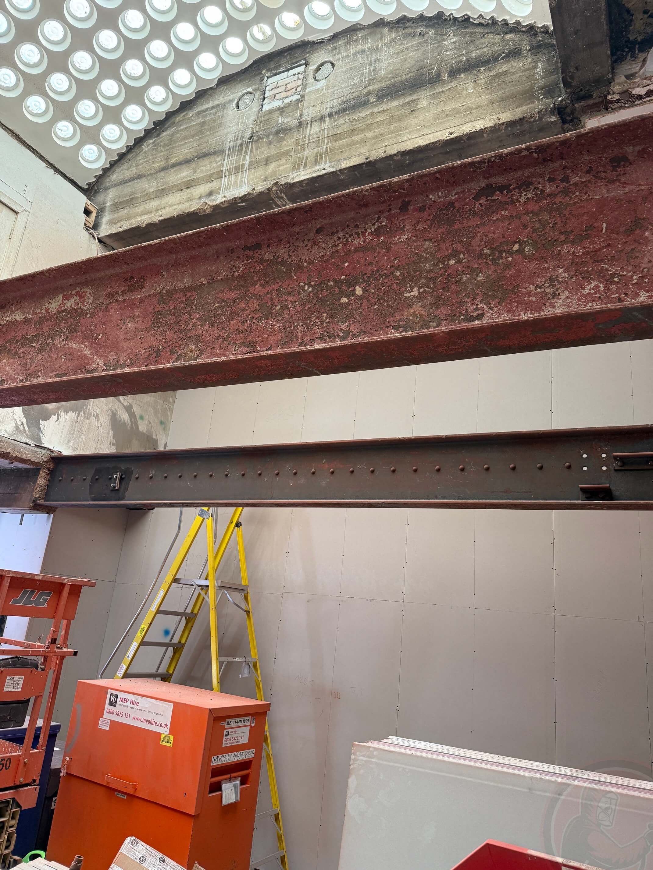 Comparison of two steel beams in construction, one coated with rust and the other cleaned, with construction equipment and yellow ladder in the background.