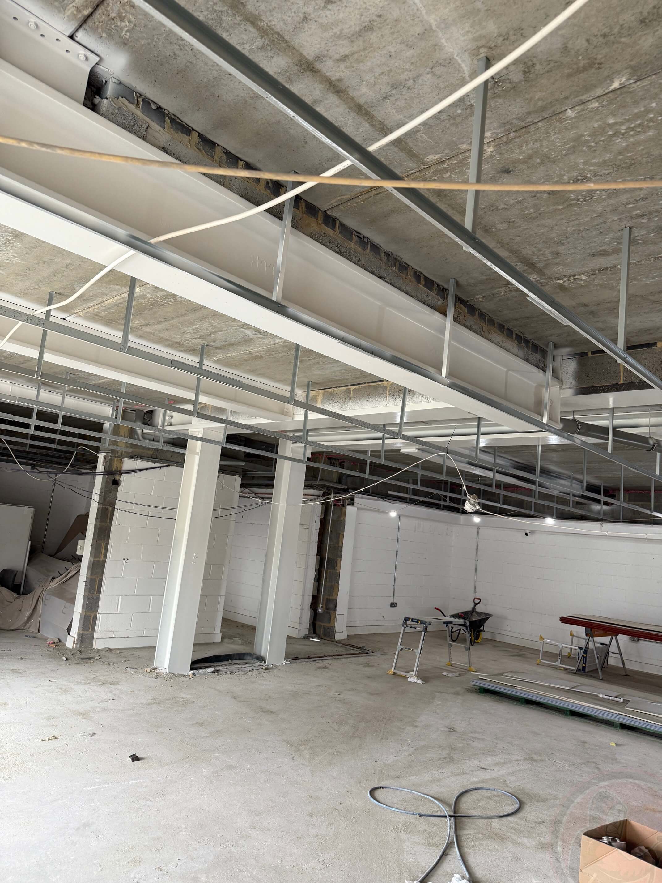 Empty unfinished industrial space with concrete floor, exposed ceiling, white painted walls, a wheelbarrow, and a workbench with materials.
