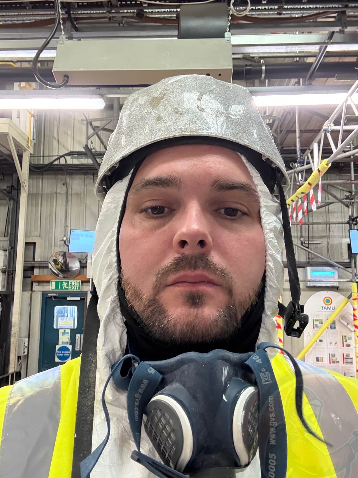 Worker wearing a safety helmet, protective suit, high-visibility vest, and a respirator mask in an industrial setting.