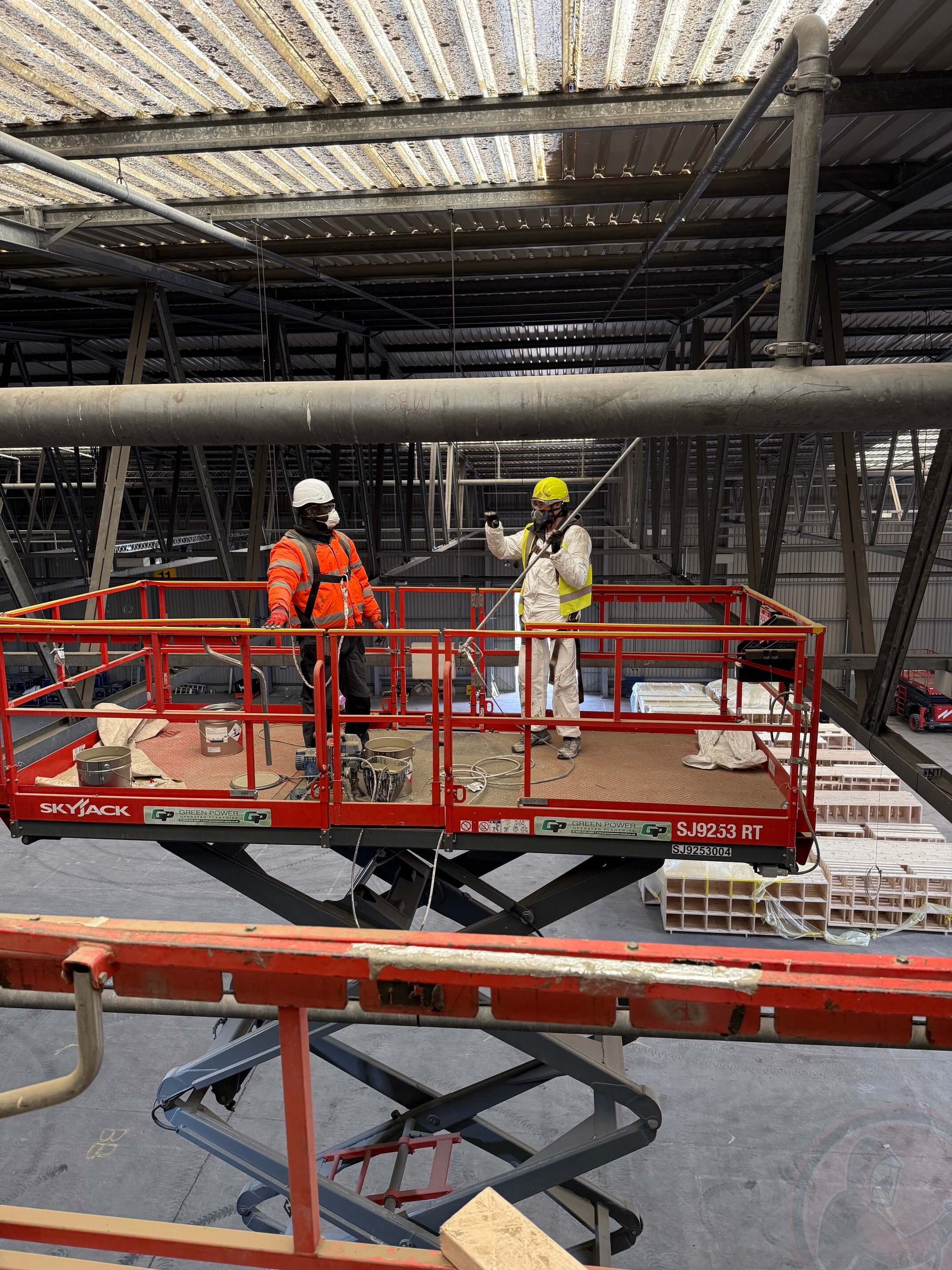 Two construction workers wearing helmets and safety gear standing on a raised red Skyjack scissor lift inside a large industrial warehouse.