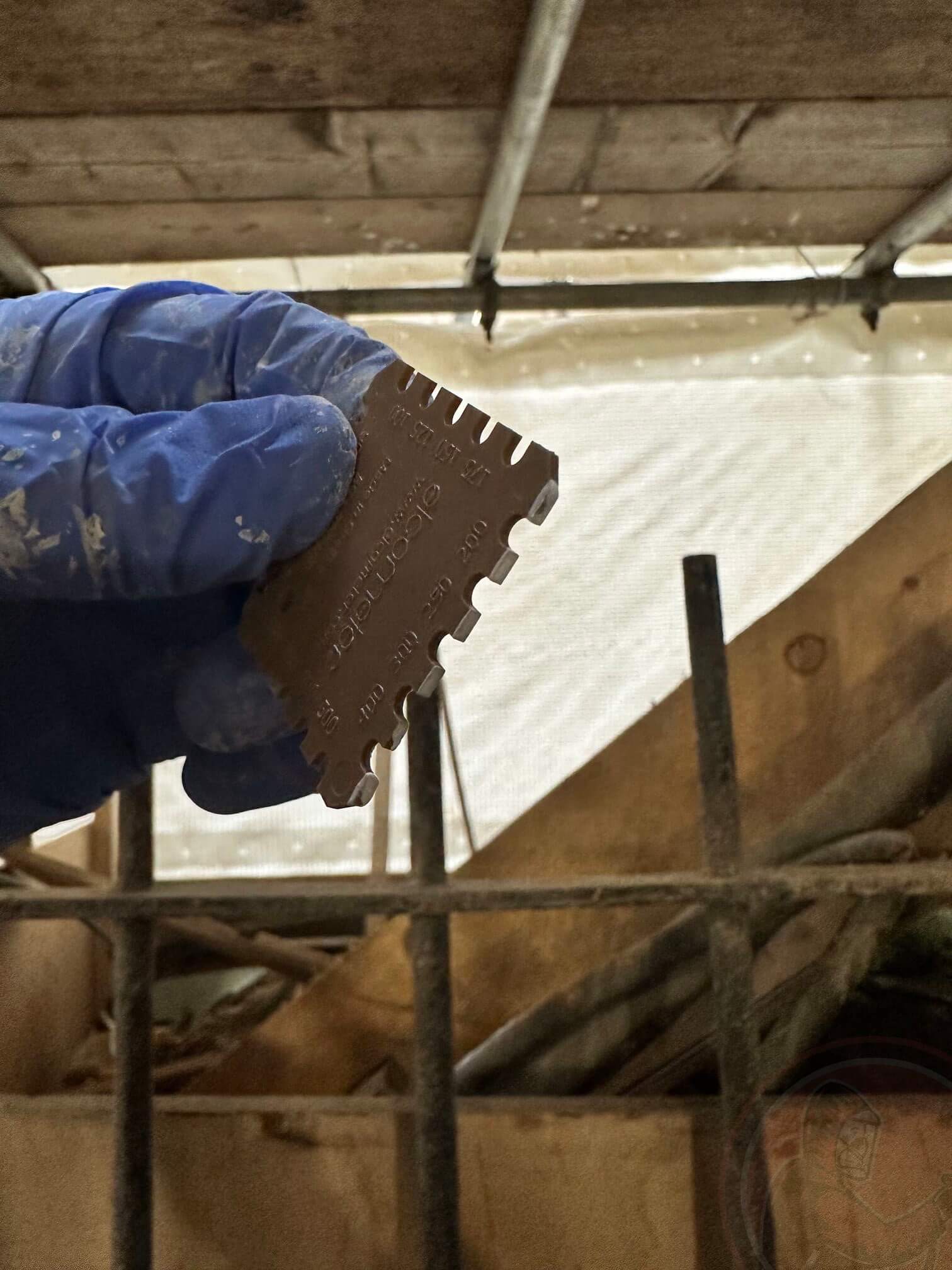 Hand wearing a blue glove holding a brown notched paint thickness gauge against a construction site background.