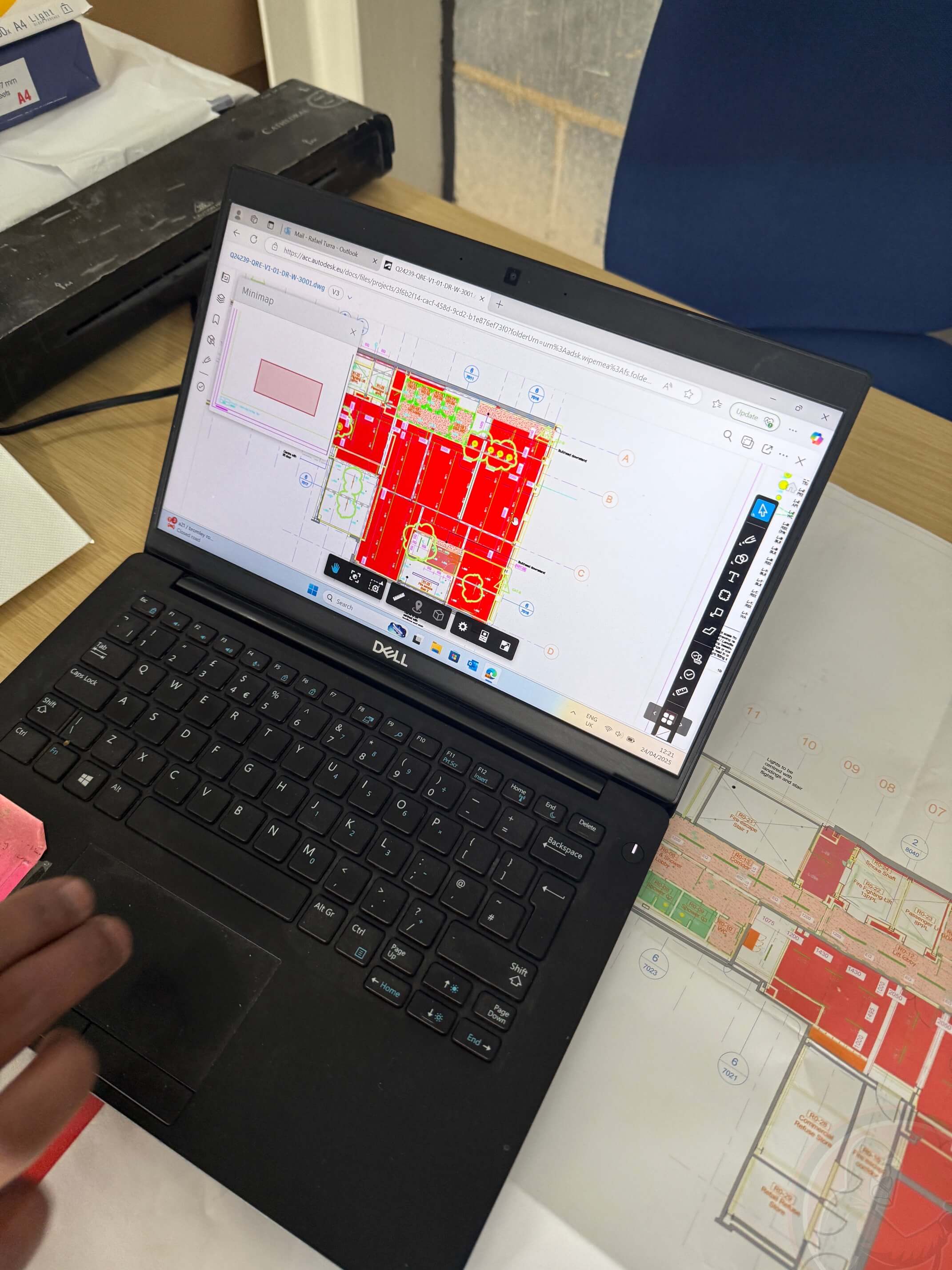 Dell laptop on a desk displaying a colorful digital construction site CAD plan with a printed site plan partially visible underneath.