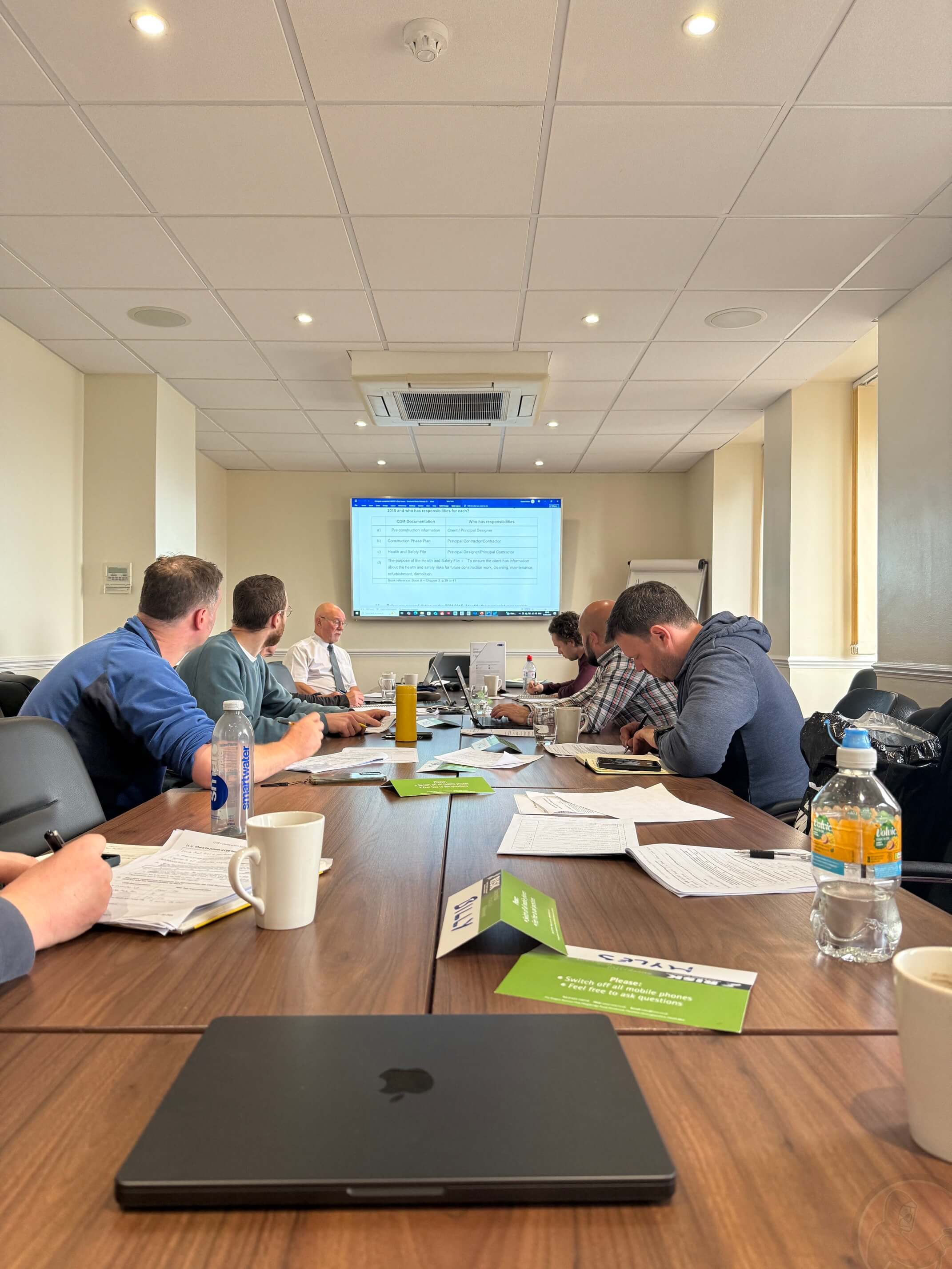 Meeting room with people seated around a table watching a presentation on a wall-mounted screen.
