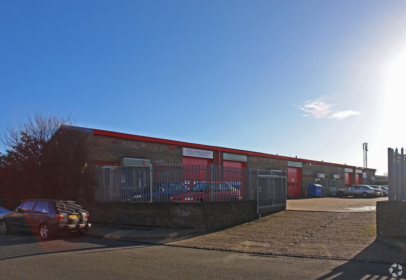 Industrial building with multiple red garage doors, surrounded by a metal fence and parked cars under a clear blue sky.