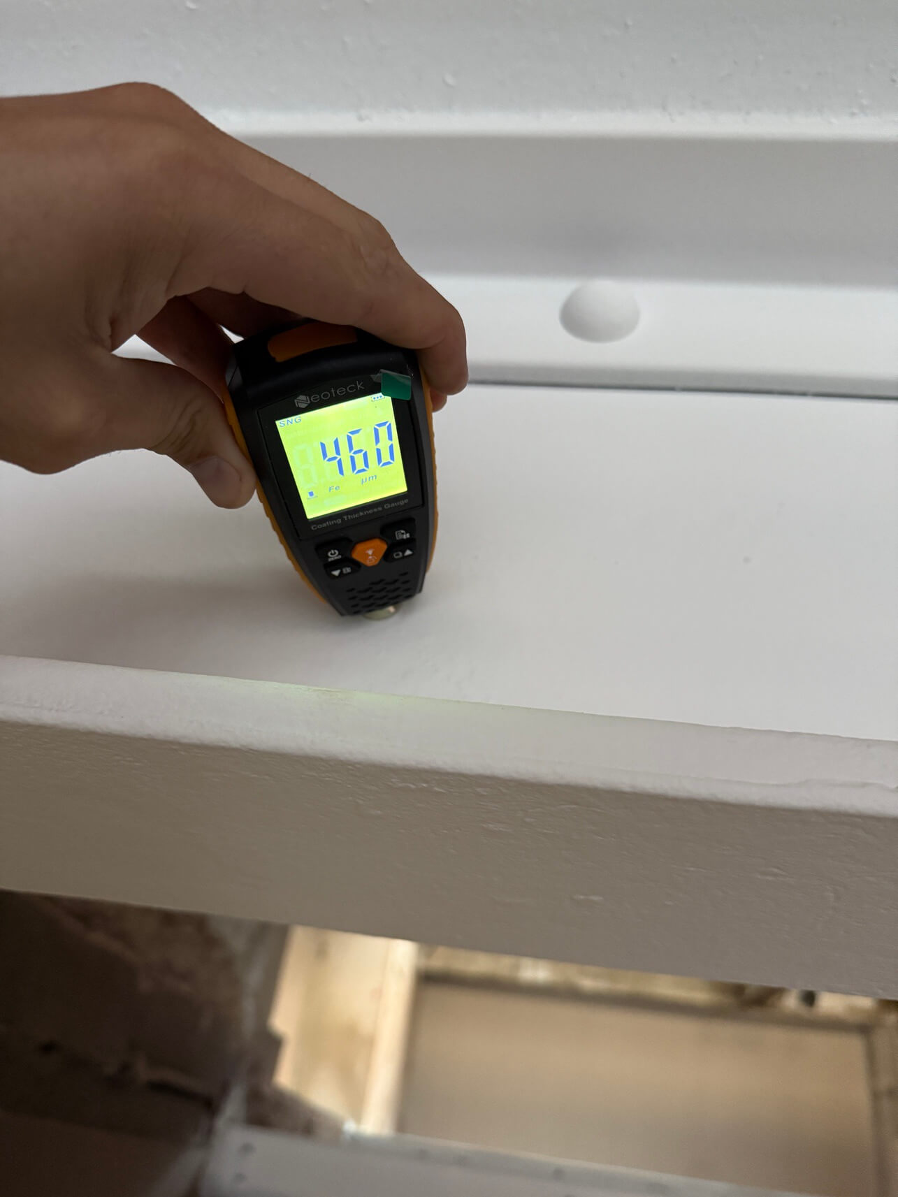 Hand holding a digital coating thickness gauge displaying a measurement of 460 micrometers on a white painted surface.