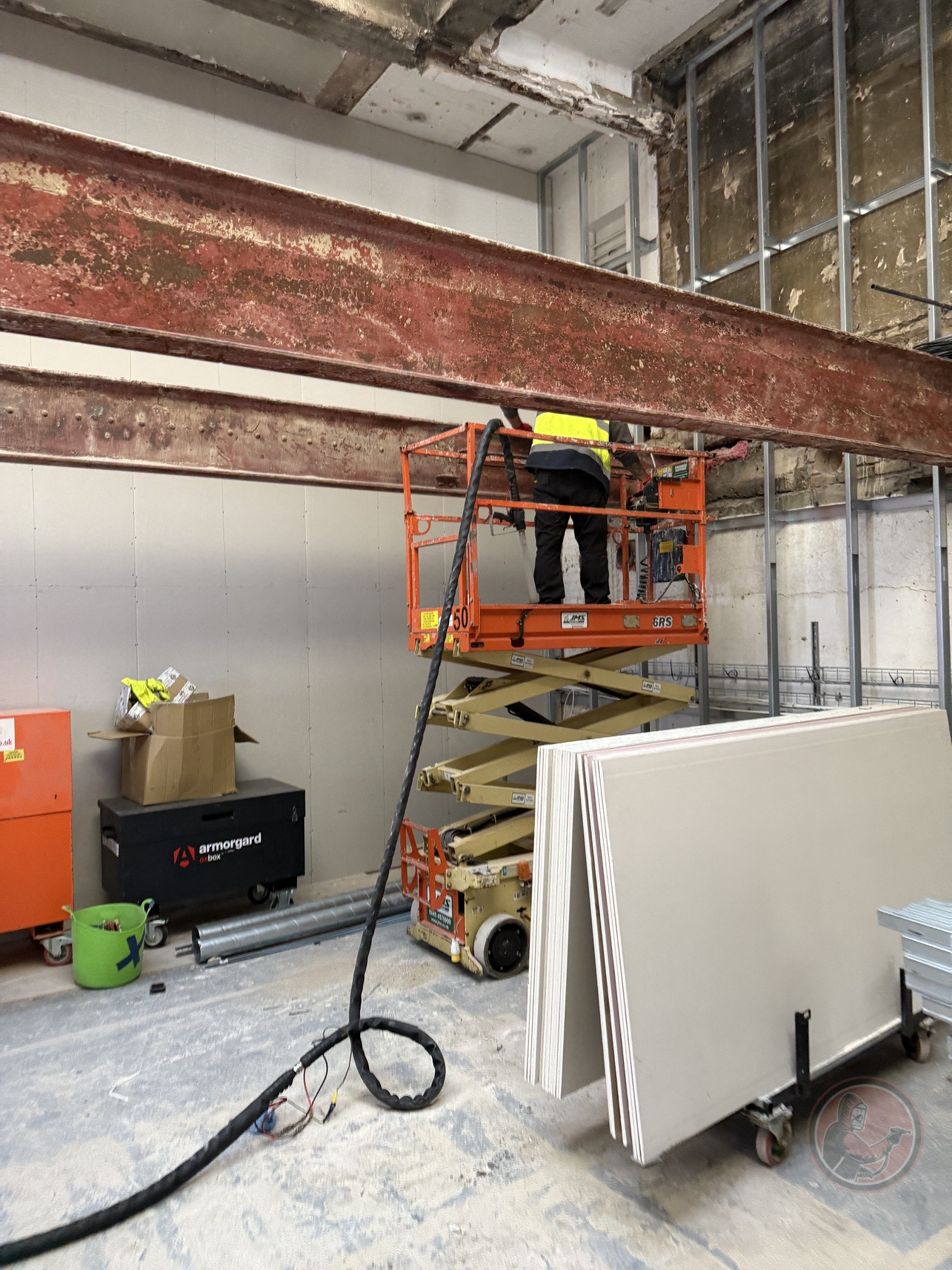Worker on an orange scissor lift dry ice blasting rusty steel beams inside a construction site with stacked drywall panels nearby.