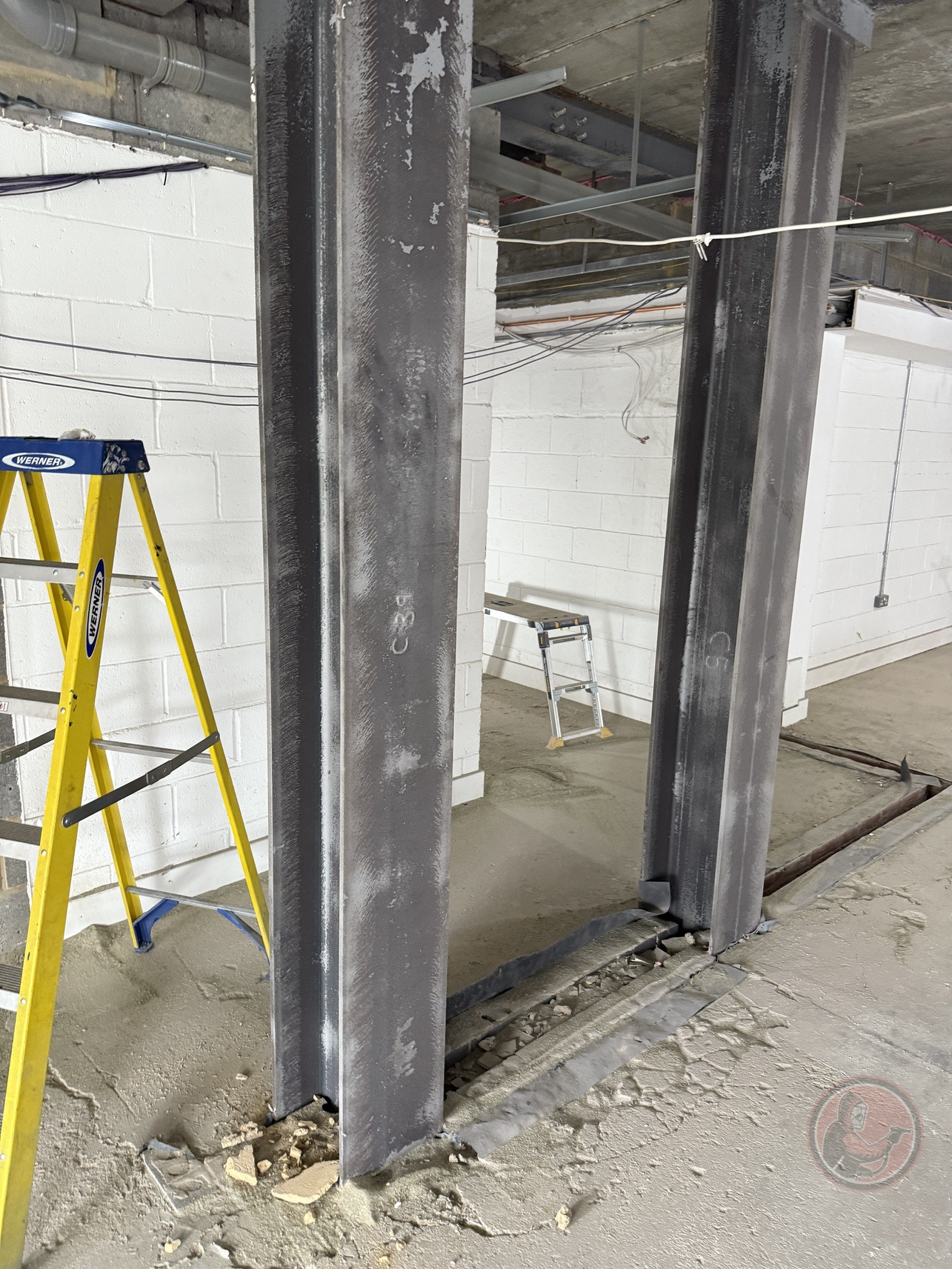 Two large vertical steel columns installed on a concrete floor with a yellow Werner ladder nearby in a construction site.
