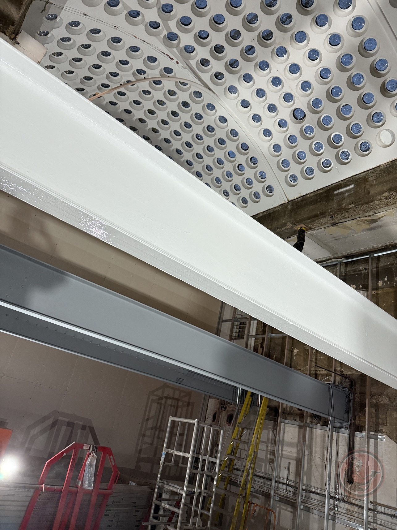 Construction site showing steel beams, scaffolding, and a ceiling with round blue-tinted light fixtures.