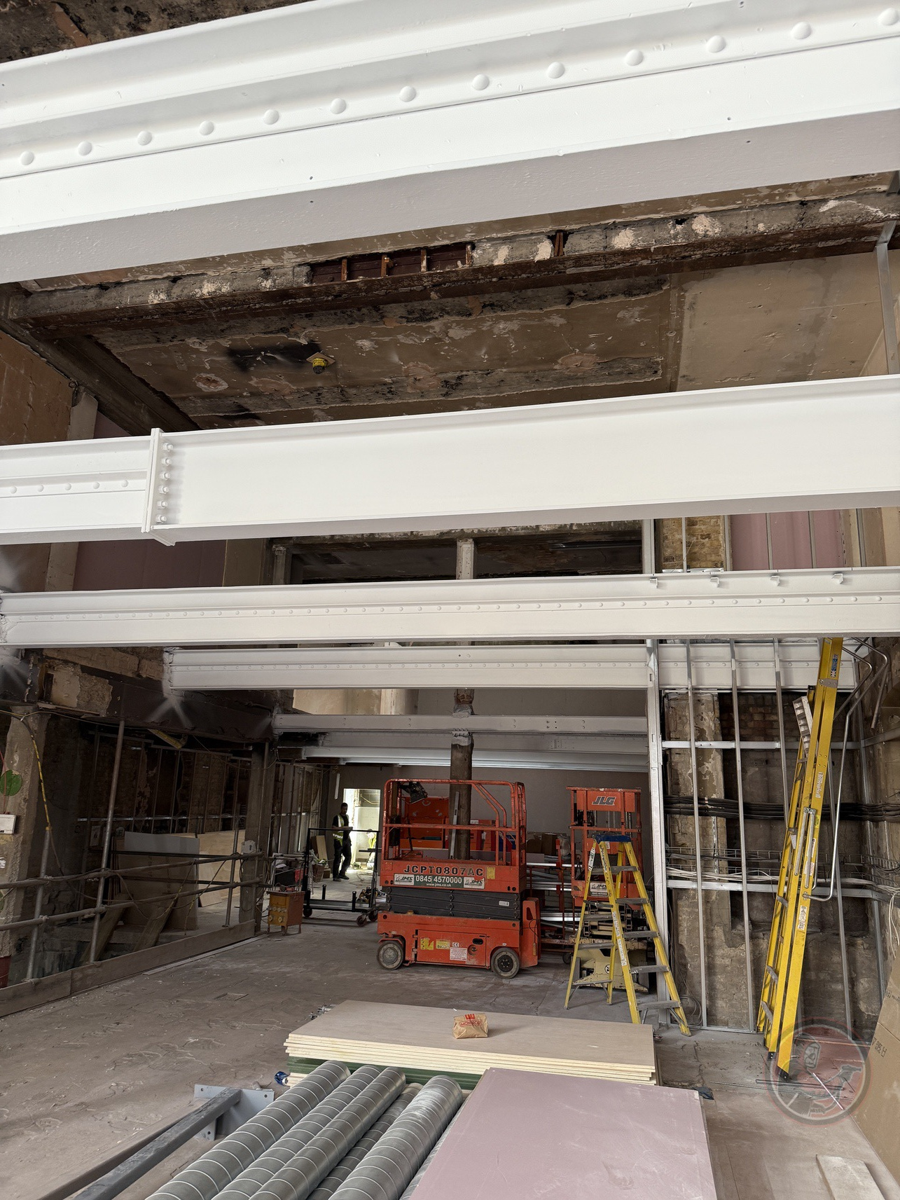 Construction site with white steel beams, orange lift, ladders, and stacked building materials.