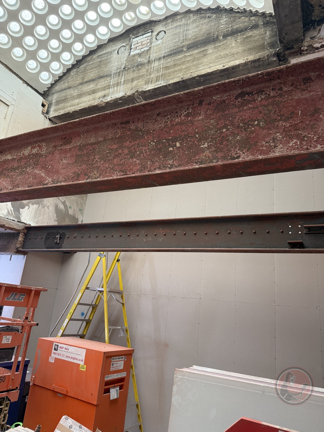 Red and gray steel beams inside construction site with yellow ladder and orange equipment.