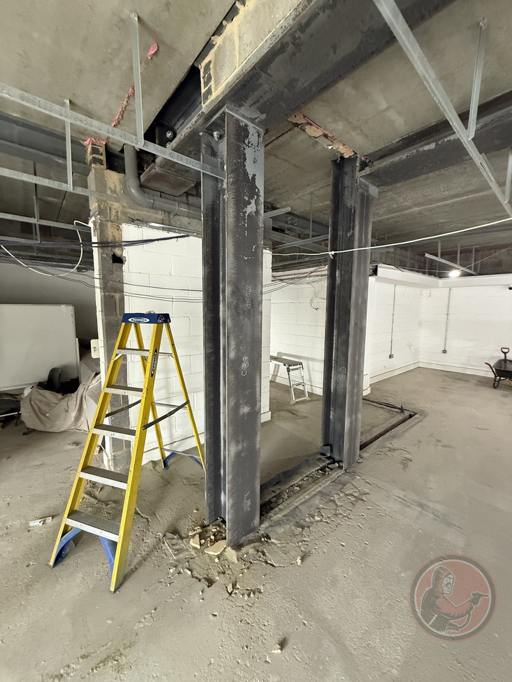 Steel columns in a construction site with a yellow ladder and exposed concrete floor and ceiling.