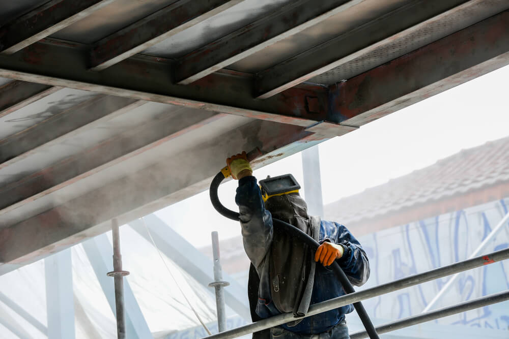 Worker wearing protective gear blasting metal structure with abrasive shot on scaffolding.