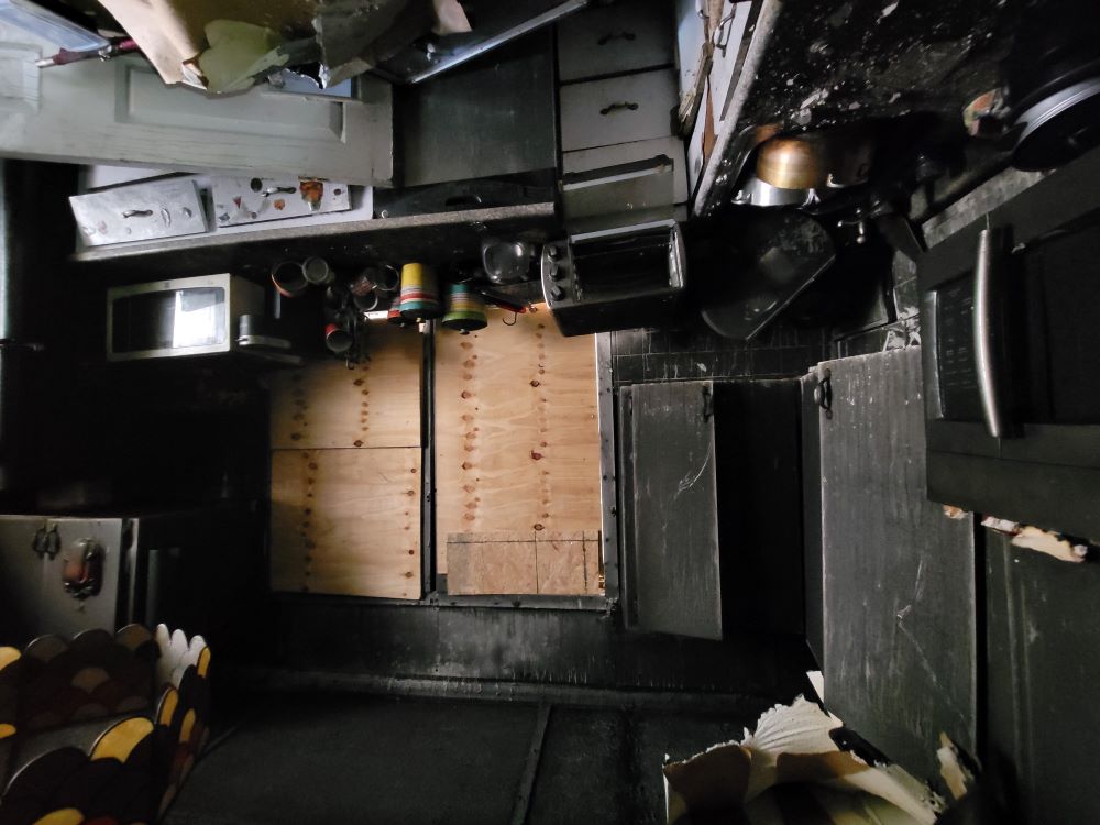 Kitchen damaged by fire with soot-covered cabinets, broken appliances, and boarded-up window.