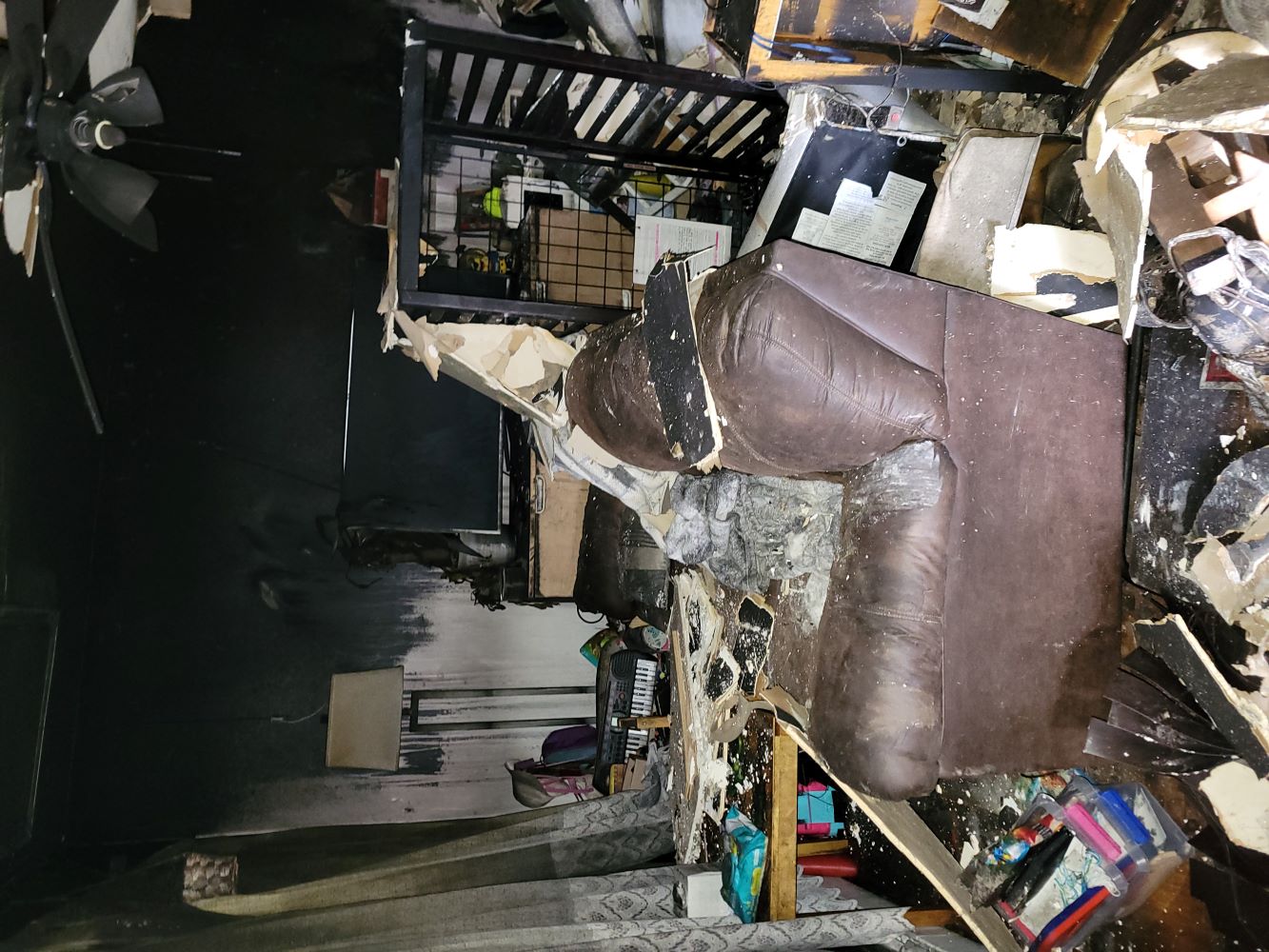 Living room heavily damaged by fire, with soot-covered walls, a damaged brown leather couch, broken ceiling materials, and debris scattered on the floor.