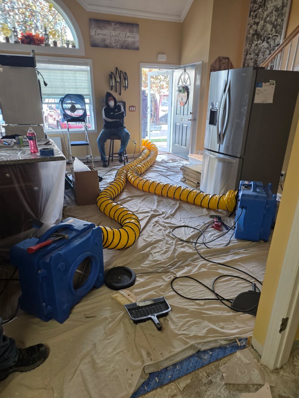 Interior of a home under renovation with protective covering on the floor, large yellow ventilation hoses connected to blue air movers, and a person wearing a mask sitting on a stool.