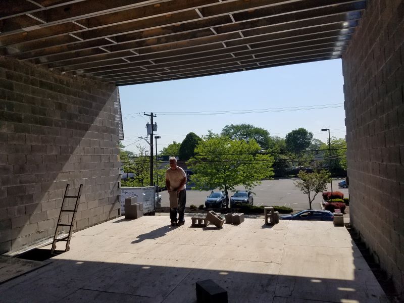Construction worker standing inside an unfinished building with concrete block walls and ceiling beams, holding a cinder block with a parking lot and trees visible outside.
