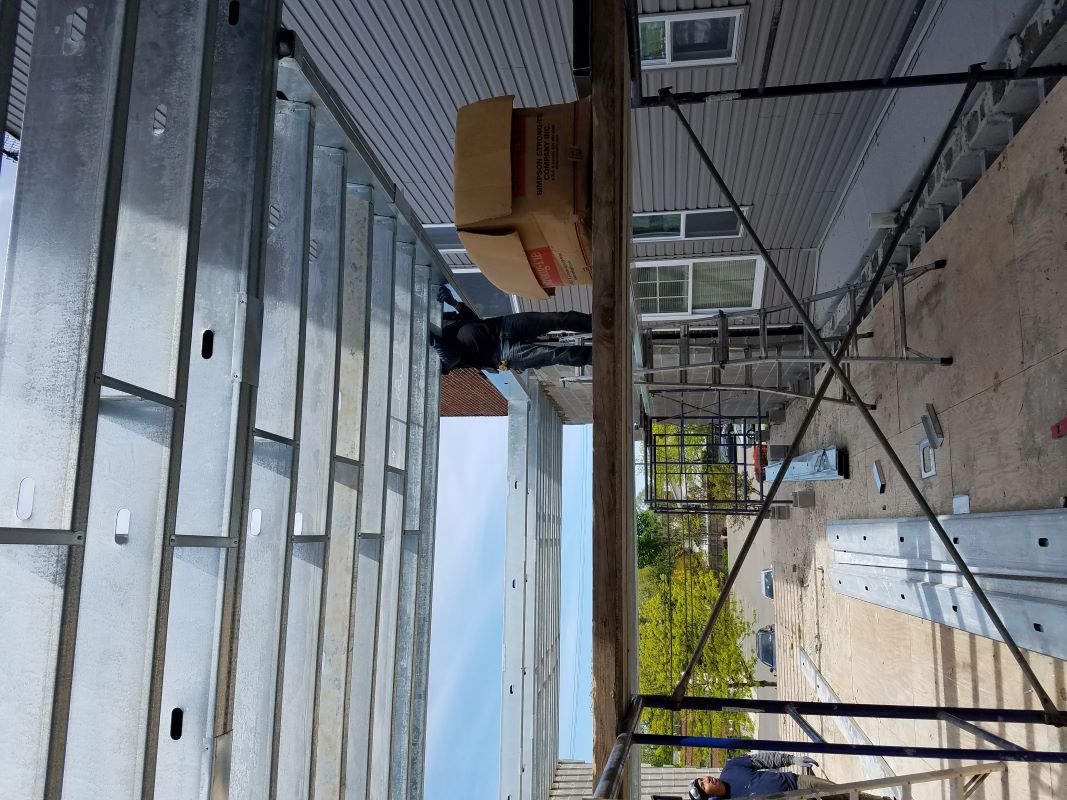 Construction workers installing metal framework on scaffolding next to a house with gray siding under a clear sky.