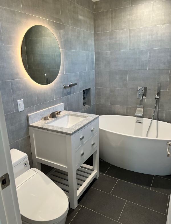 Modern bathroom with gray tiled walls and floor, featuring a white freestanding bathtub, white vanity with marble countertop, round backlit mirror, and a toilet.