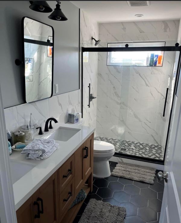 Modern bathroom with black hexagonal tile floor, wooden vanity with white countertop, rectangular mirror, and glass shower enclosure with marble walls.