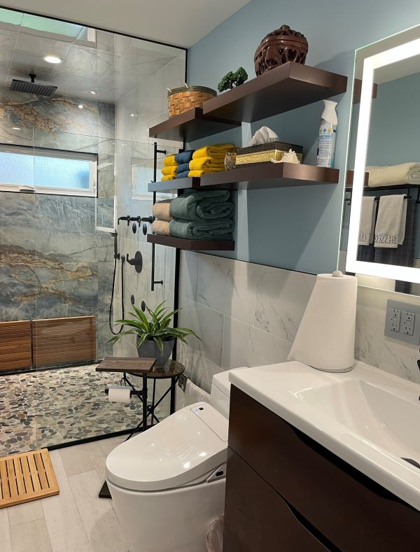 Modern bathroom with glass-enclosed stone-tiled shower, white toilet, wooden shelves holding folded towels and decor, and illuminated mirror above sink.