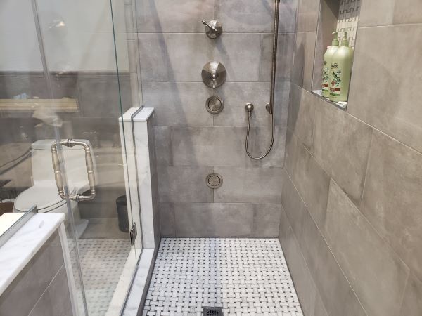 Modern walk-in shower with gray tile walls, mosaic tile floor, stainless steel shower fixtures, glass door, and shampoo bottles in a wall niche.