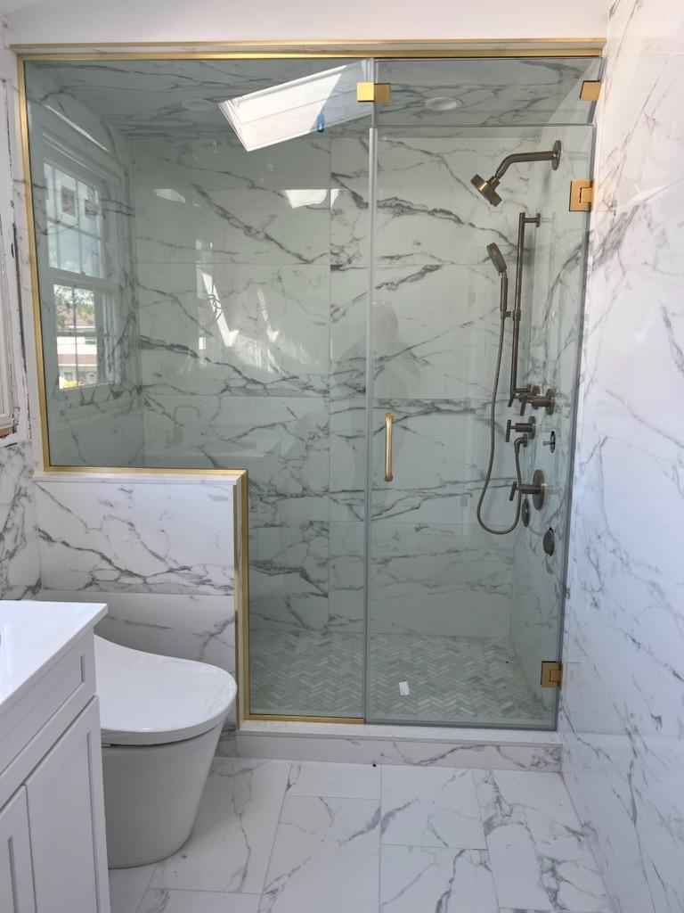 Modern bathroom with white marble tiles, a glass shower enclosure with gold trim, and bronze shower fixtures.