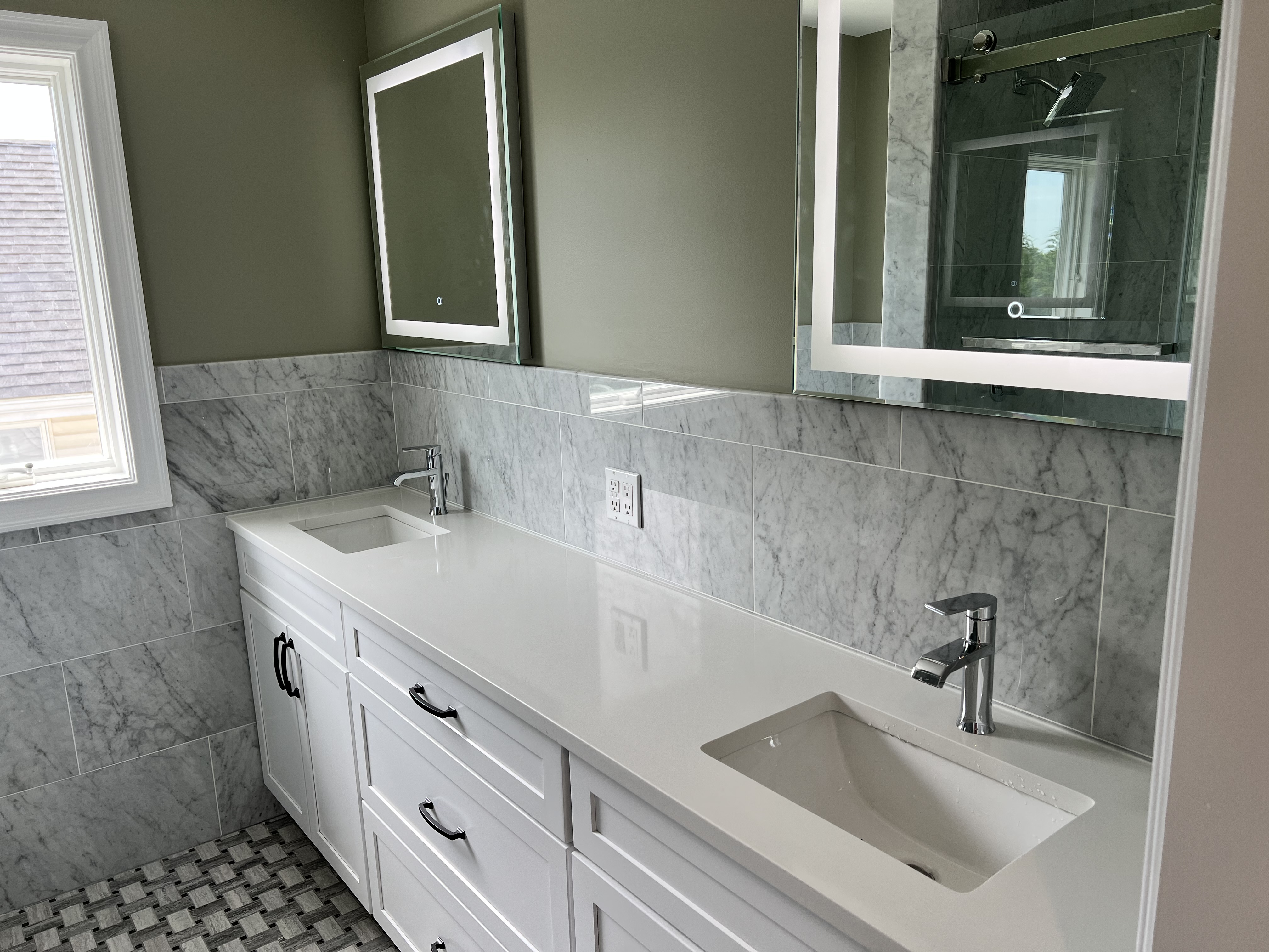 Modern bathroom vanity with white double sinks, chrome faucets, illuminated mirrors, and marble tiled walls and floor.