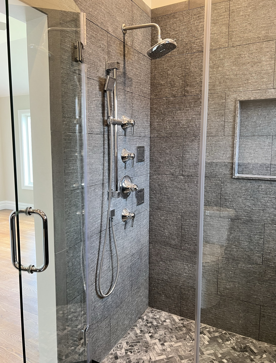 Modern shower with textured gray wall tiles, chrome showerhead, handheld shower, multiple controls, and glass door.