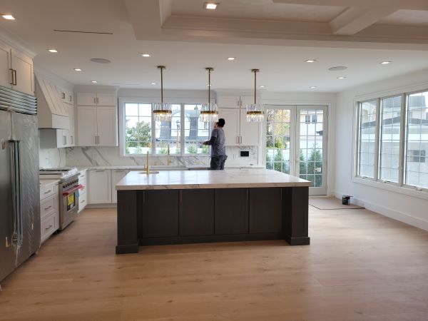Modern kitchen with large dark island, white cabinets, marble countertops, hardwood floor, and a person standing near the window.