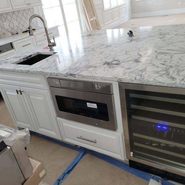 Modern kitchen island with white cabinetry, marble countertop, built-in stainless steel microwave, small sink with faucet, and a wine cooler with digital display.