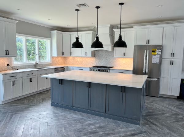 Modern kitchen with white cabinetry, gray island with white countertop, three black hanging pendant lights, stainless steel refrigerator, and large window.