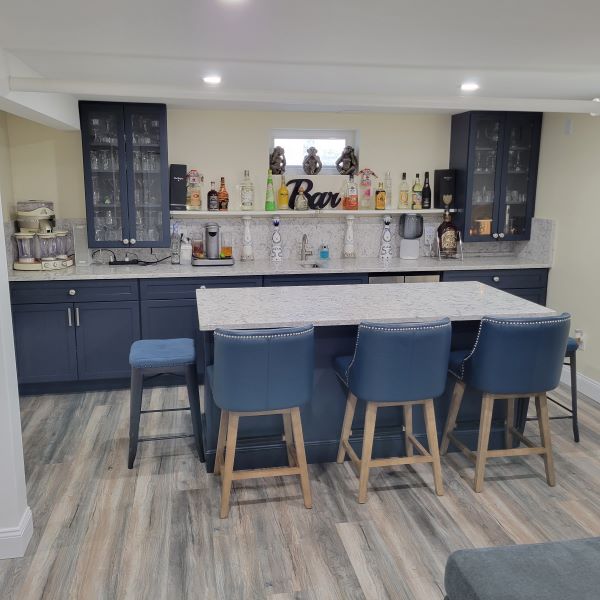 Home bar setup with navy blue cabinets, a marble countertop island with four blue bar stools, and various bottles and glassware on the counter and in glass-paneled cabinets.
