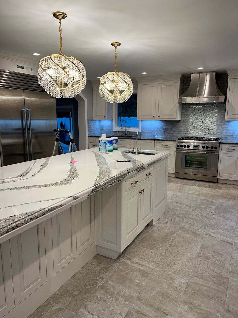Modern kitchen with white marble island, crystal pendant lights, stainless steel appliances, and tiled floor.