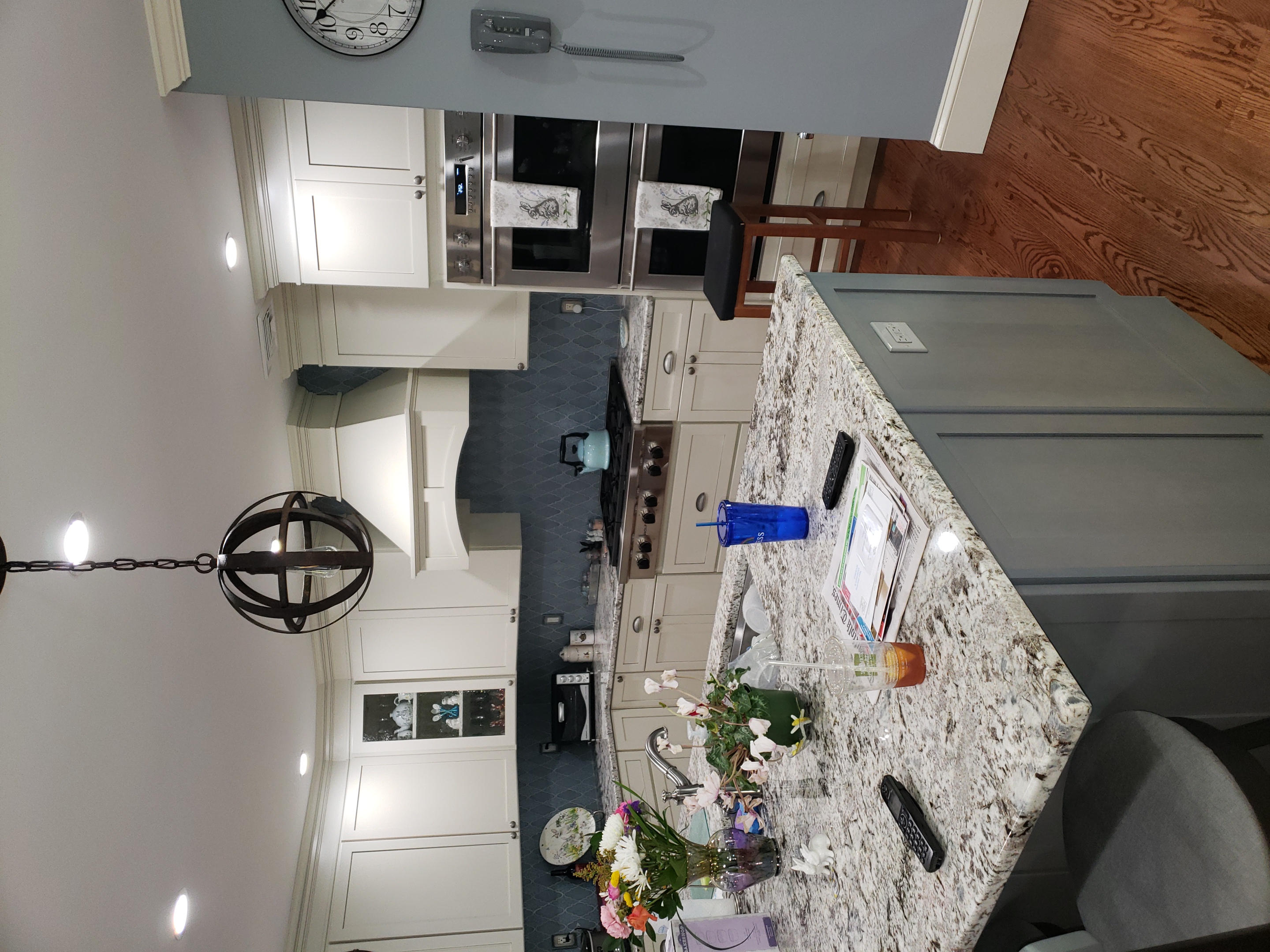 Modern kitchen with light-colored cabinets, granite countertop island, stainless steel double ovens, and hanging round pendant light.