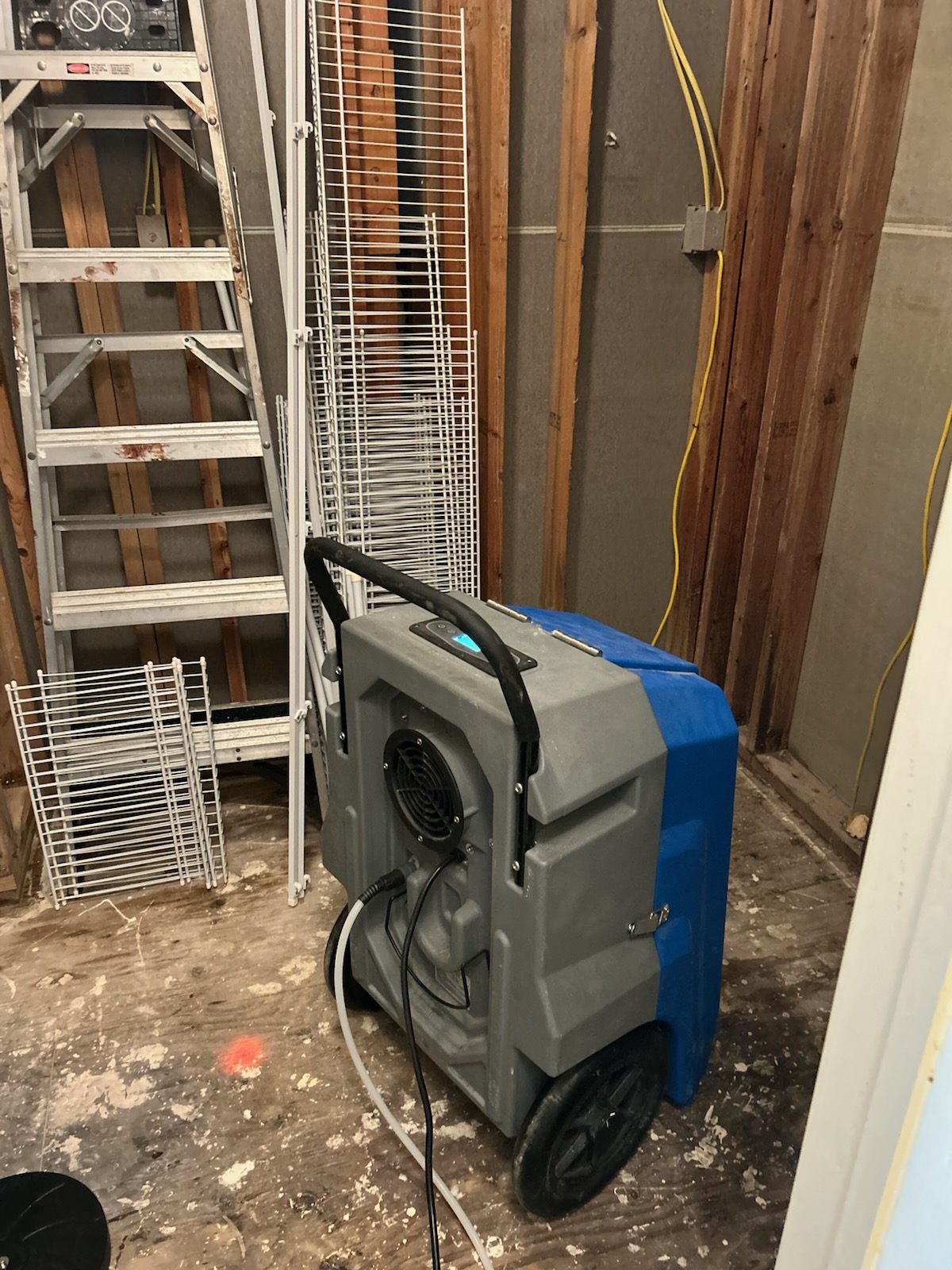 Large industrial gray and blue dehumidifier with hoses and cables in an unfinished room with exposed wooden studs and a metal ladder.