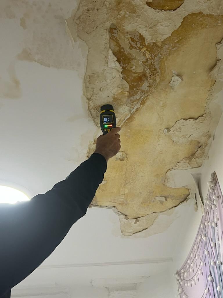 Hand holding a moisture meter against a large water-damaged and peeling ceiling.