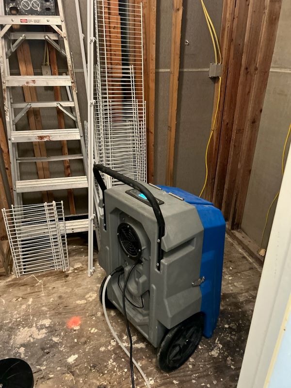Dehumidifier with a gray and blue body on wheels inside a wooden framed room under construction with a metal ladder and white racks leaning against the wall.