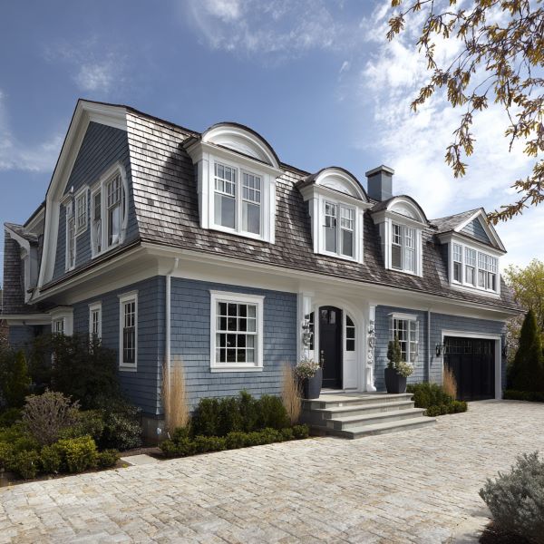 Two-story blue house with white trim, dormer windows, a stone driveway, and greenery in front under a partly cloudy sky.