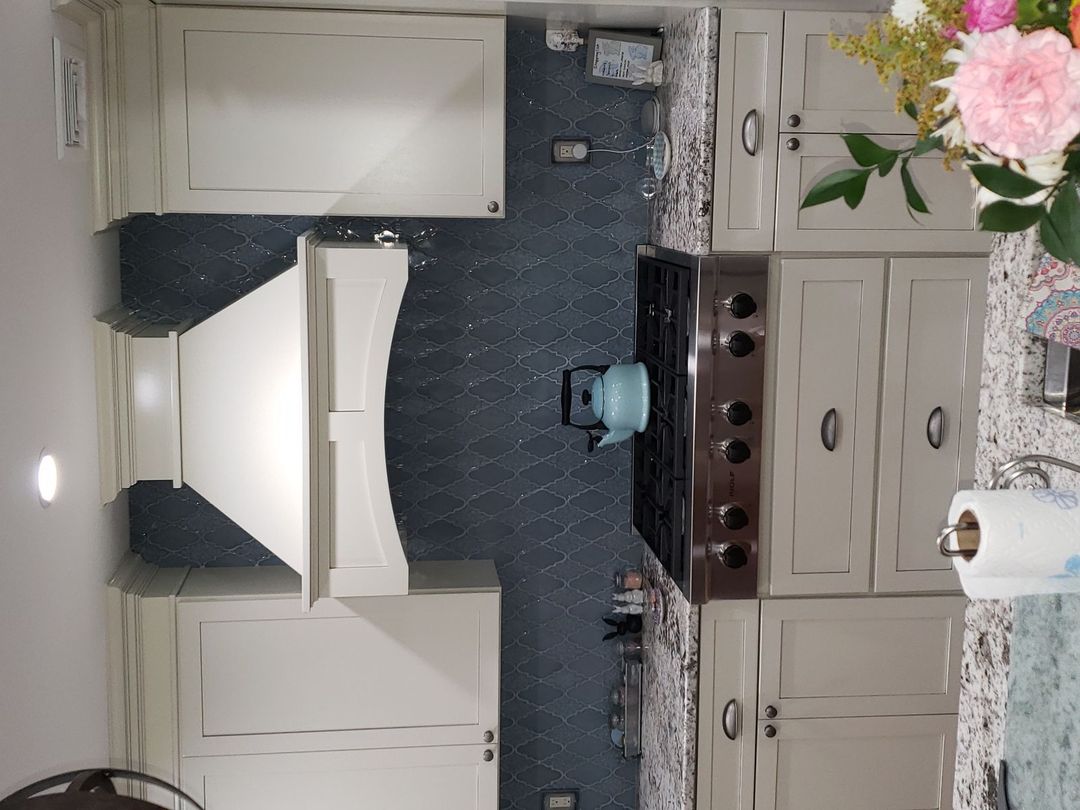 Modern kitchen countertop with granite surface, white cabinets, a stainless steel stove with black knobs, a light blue kettle, and a white range hood against a blue tile backsplash.