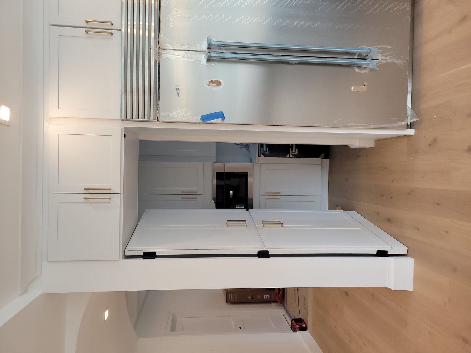 Modern kitchen corner with white cabinets, built-in microwave, and a large stainless steel refrigerator partially covered in protective wrap.