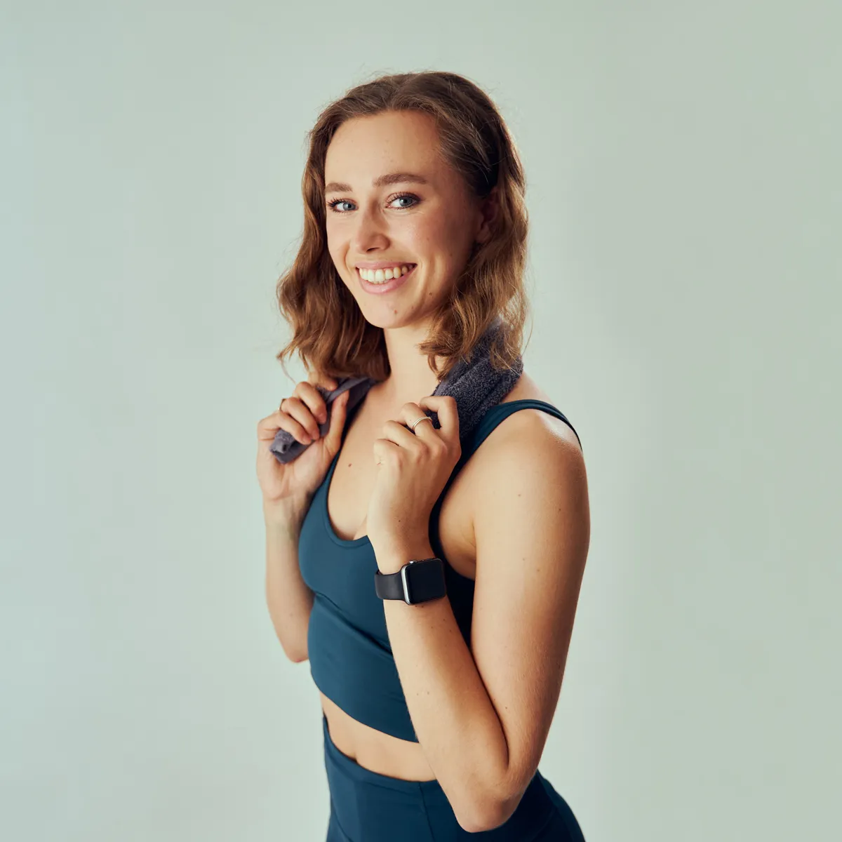 Smiling woman in dark athletic wear holding a towel around her neck with a smartwatch on her wrist.