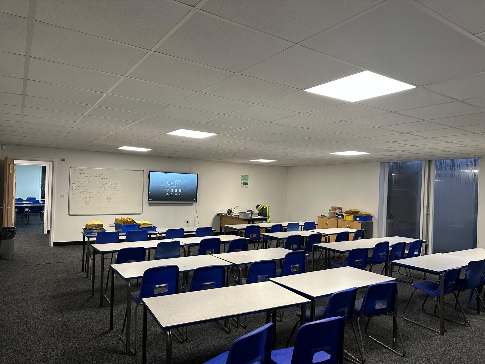 Empty classroom with blue chairs and white desks, whiteboard with handwritten notes, a wall-mounted TV screen, and large windows on the right.