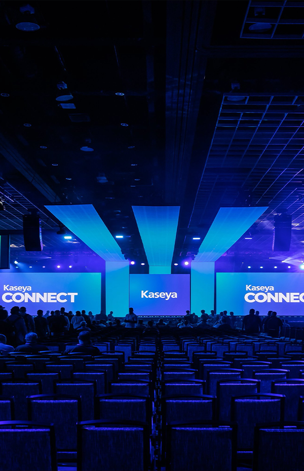 Large conference room with rows of empty chairs facing three large screens displaying Kaseya Connect branding under blue and purple lighting.