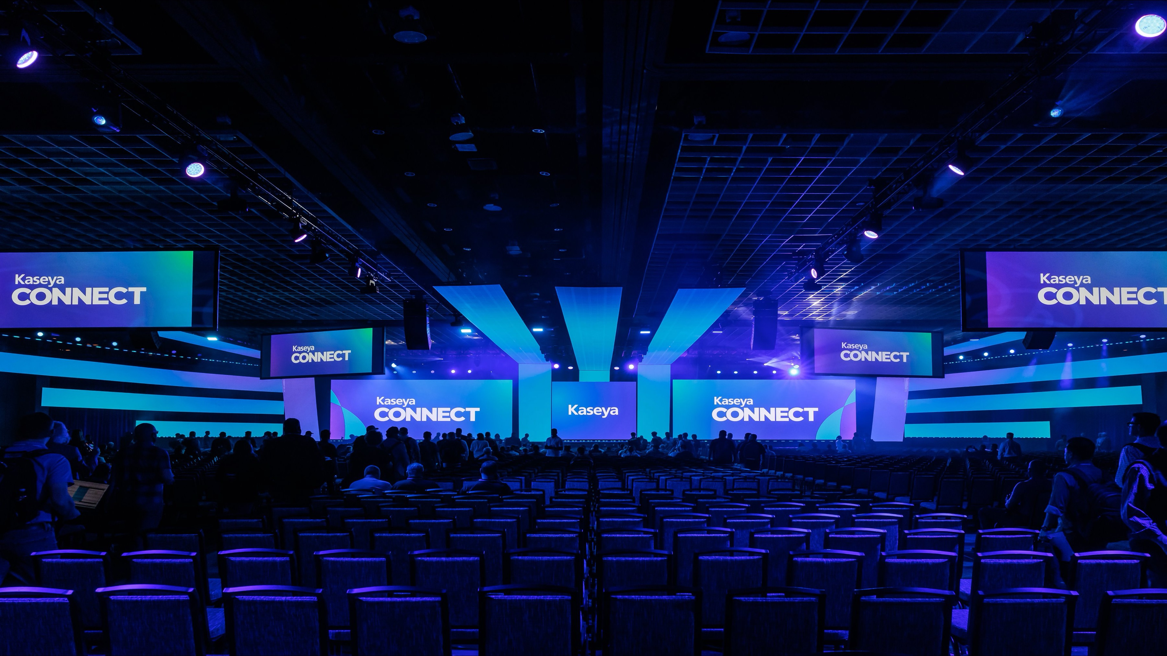 Conference hall with rows of chairs and multiple large screens displaying 'Kaseya CONNECT' in blue lighting.