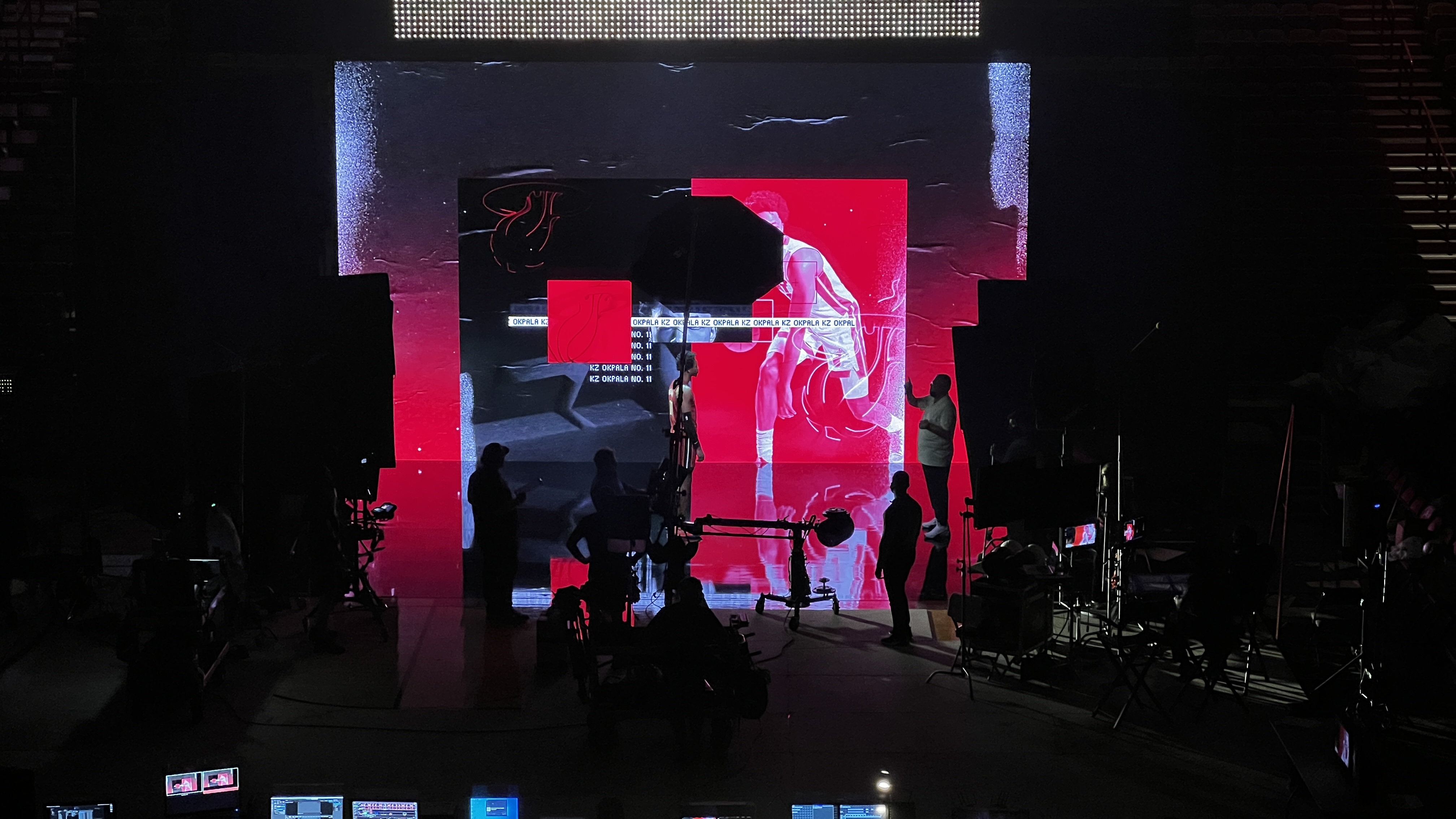 Dark studio with silhouettes of crew members and filming equipment in front of a large LED screen displaying a basketball player in white uniform on a red and black background.